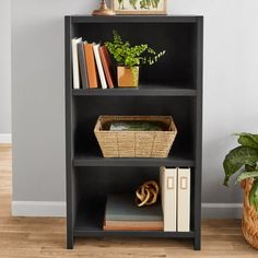 a black bookcase with books, plants and pictures on it in front of a gray wall