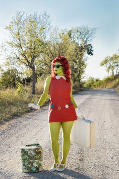 a woman in green makeup holding a suitcase and walking down a dirt road with trees behind her