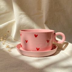 a pink cup and saucer with red hearts on it sitting on a white cloth