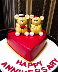 two teddy bears sitting on top of a heart shaped cake with the words happy anniversary