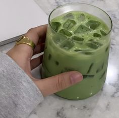 a hand holding a green drink with ice cubes in it on a marble table