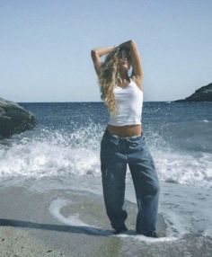 a woman standing on top of a sandy beach next to the ocean with her arms behind her head