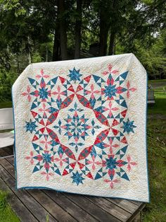 a quilt is sitting on a wooden table outside in the grass with trees behind it