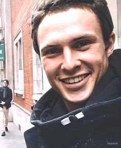 a man smiling while standing in front of a brick building on a city street with people walking by