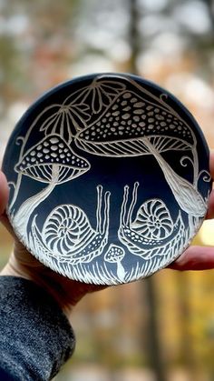 a hand holding a black and white bowl with designs on it's surface in front of trees