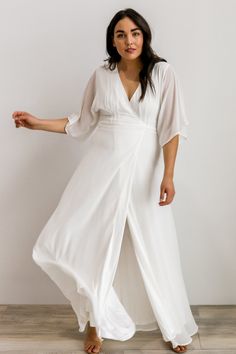 a woman wearing a white dress and sandals standing in front of a wall with her arms out