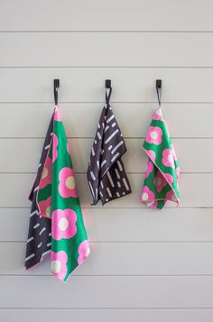 three towels hanging from hooks on a wall with white siding behind them and pink flowers in the center