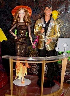 two dolls are standing on a table in front of a wallpapered room with gold foil