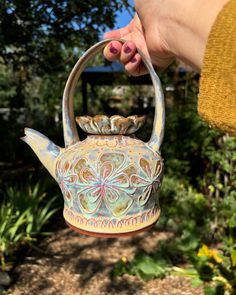 a hand holding a teapot with an intricate design on it's handle, in front of some trees