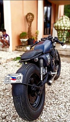 a black motorcycle parked on top of a gravel covered ground next to a building with people sitting in the background