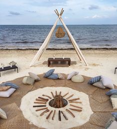 an outdoor fire pit on the beach with pillows and blankets around it, in front of the ocean