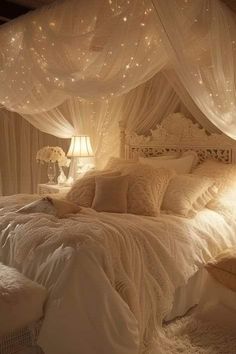 a white bed covered in lots of pillows and blankets next to a night stand with lights on it