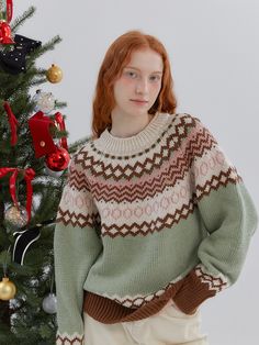 a woman standing in front of a christmas tree wearing a green sweater and white pants