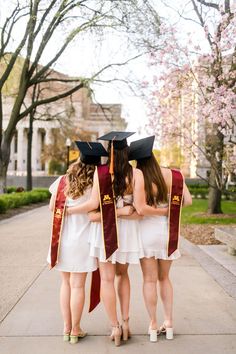 three young women in graduation gowns are standing on the sidewalk with their backs to each other