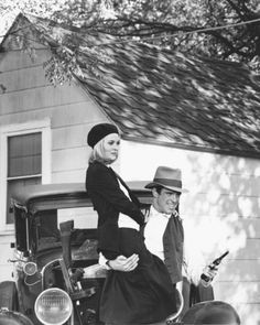 a man and woman standing next to an old car in front of a house with a dog