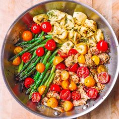 a pan filled with chicken, tomatoes and asparagus on top of a wooden table