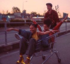 three people sitting in a shopping cart on the side of the road with their arms up