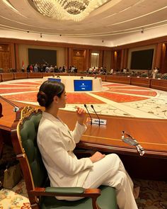 a woman sitting at a table in front of a boardroom full of people with papers