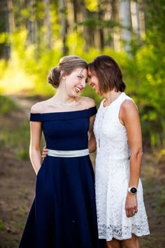 two women standing next to each other on a dirt road in the woods with trees behind them