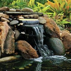 a small waterfall made out of rocks and stones in a pond surrounded by greenery