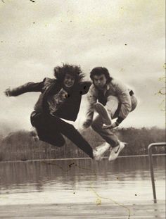 two people jumping in the air on a skateboard over a body of water with trees behind them