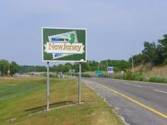 a welcome to new jersey sign on the side of an empty road with grass and trees in the background