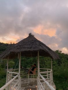 a person sitting on a bench under a thatched roof in the middle of a forest