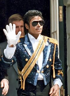 a man in a suit and sunglasses waves to the crowd from an open door with his hand up