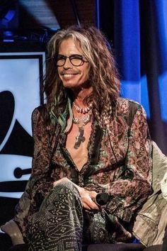 a man with long hair and glasses sitting on a chair in front of a microphone