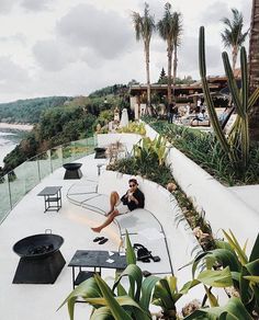 a woman sitting on the edge of a swimming pool next to plants and chairs with an ocean view in the background