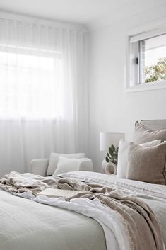 an unmade bed in a bedroom with white walls and curtains on the windowsill