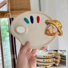 a person holding up a plate with different colored objects on it and the image of a man's face painted on it