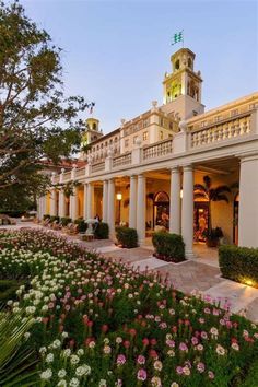 a large white building with columns and flowers in the front yard at night time, surrounded by greenery