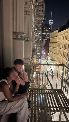 a man and woman sitting on a balcony looking out at the city lights in the distance