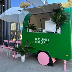 a green food truck parked on the side of a street next to pink chairs and umbrellas