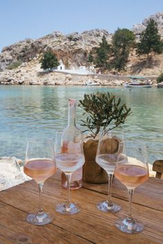 three wine glasses sitting on top of a wooden table next to the ocean and mountains