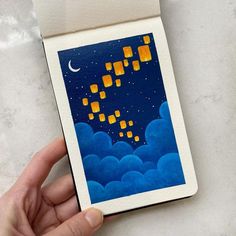 a hand is holding up a small card with some paper lanterns floating in the sky