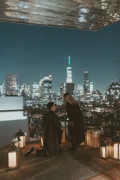 two women standing on top of a building looking out at the city lights and skyscrapers