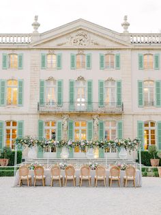 a long table with chairs and flowers in front of a large white building that has green shutters