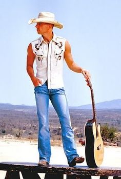 a man standing on top of a wooden sign with a guitar in his hand and wearing a cowboy hat