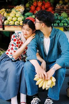 a man and woman sitting next to each other in front of fruit stand with bananas