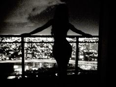 a woman standing on top of a balcony next to a window at night with city lights in the background