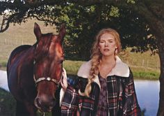 a woman standing next to a brown horse near a tree and water in the background