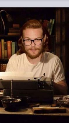 a man sitting at a table with a typewriter in front of him and books on the shelves behind him