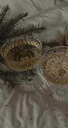 two wine glasses sitting on top of a bed next to a pine branch and candle