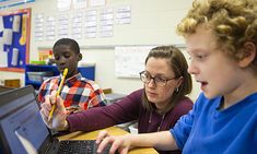 two children and an adult are working on laptops in a classroom with other students