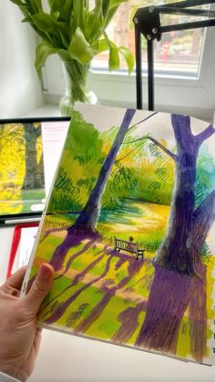 a person holding up a drawing in front of a window with trees and bench on it