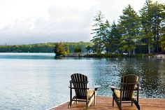 two chairs sitting on a wooden dock next to a body of water with trees in the background