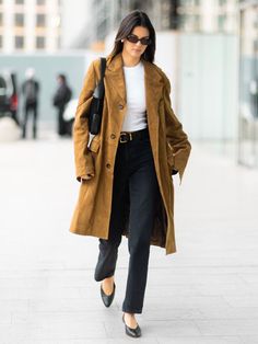 a woman walking down the street wearing a brown coat and black pants with her hand in her pocket