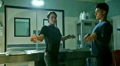 two men standing in a kitchen talking to each other and one is pointing at something